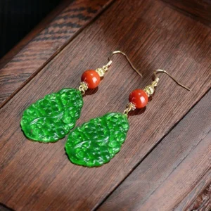 Close-up side view of carved green jade drop earrings with red agate top bead and gold-toned sterling silver posts on wooden surface