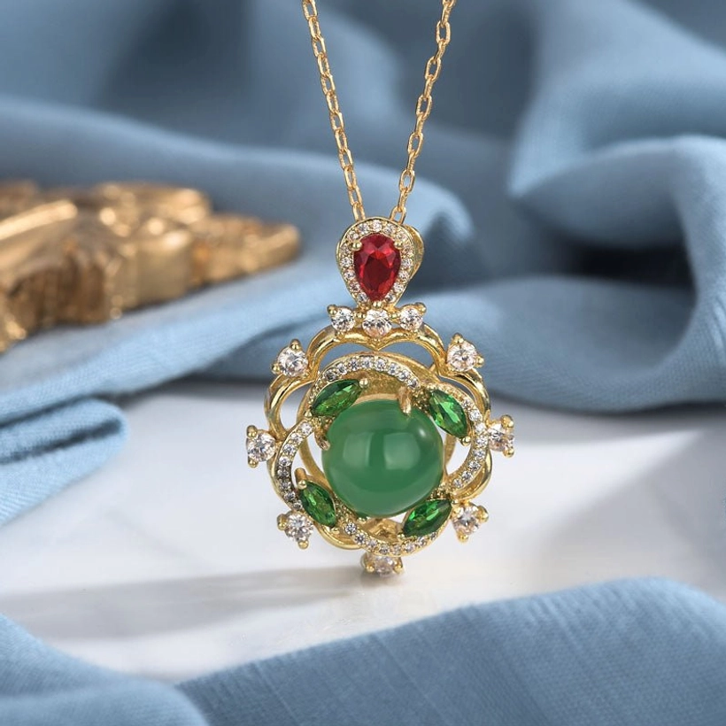 Green jade and red crystal pendant necklace on blue fabric, showing ornate gold setting