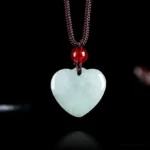 Pale green heart-shaped jade pendant necklace with brown braided cord and red accent bead, frontal view on black background