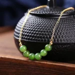 14K gold-plated Hetian cyan jade beaded necklace displayed on wooden tray with traditional teapot, showcasing delicate gold chain and arc-shaped arrangement of seven green jade beads - prosperity and harmony luck necklace