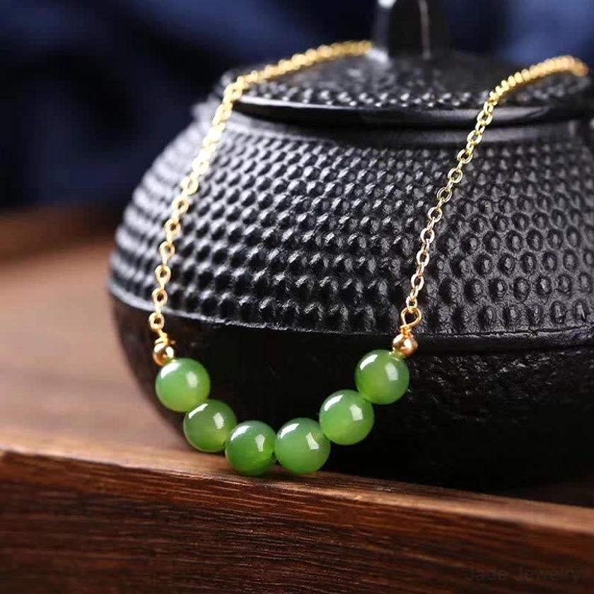 14K gold-plated Hetian cyan jade beaded necklace displayed on wooden tray with traditional teapot, showcasing delicate gold chain and arc-shaped arrangement of seven green jade beads - prosperity and harmony luck necklace