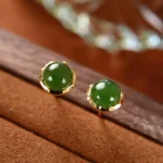 Hetian cyan jade stud earrings with gold-plated setting displayed on brown velvet, showing intricate floral bezel design and natural jade beads