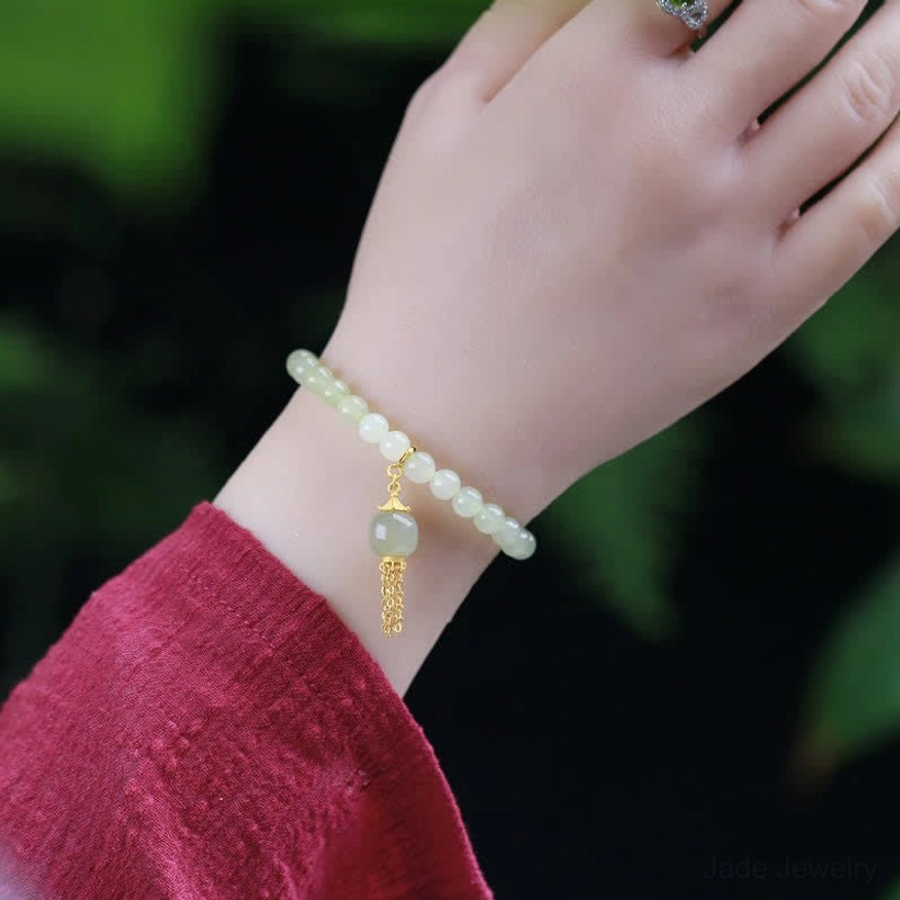 Hetian jade bracelet worn on wrist with red traditional silk sleeve, delicate jade bead stack with gold tassel pendant close-up