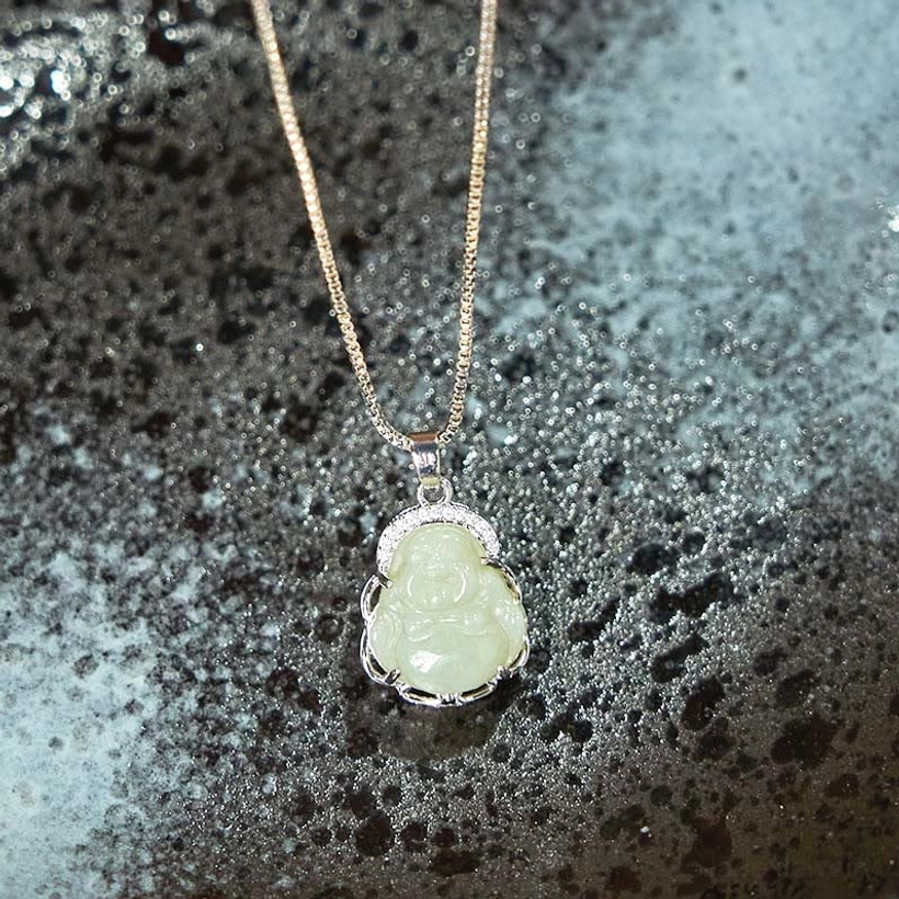 Light green jade Laughing Buddha pendant with round rhinestone frame and silver chain, flat lay on dark ceramic