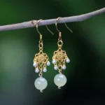Hetian jade flower dangle earrings with 14K gold plating and light green jade drops, displayed on natural green twig with blurred outdoor background