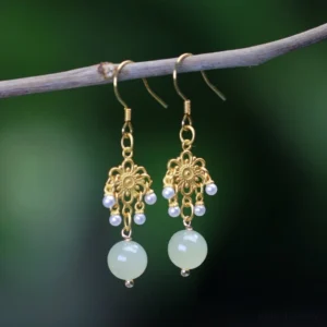 Hetian jade flower dangle earrings with 14K gold plating and light green jade drops, displayed on natural green twig with blurred outdoor background
