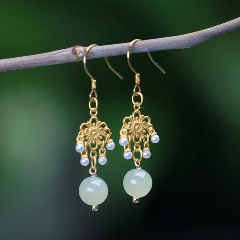 Hetian jade flower dangle earrings with 14K gold plating and light green jade drops, displayed on natural green twig with blurred outdoor background