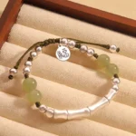 Hetian jade Fu character bamboo bracelet with sterling silver beads on dark green woven cord, flat lay display on cushion