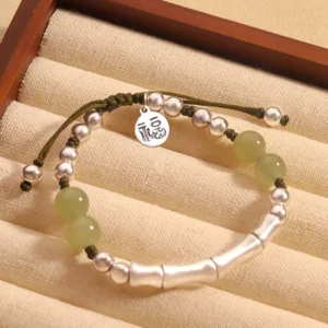 Hetian jade Fu character bamboo bracelet with sterling silver beads on dark green woven cord, flat lay display on cushion