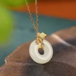 Hetian jade ginkgo leaf peace buckle necklace pendant on brown leaf tray, showing polished white nephrite jade with gold-plated ginkgo bail and delicate chain