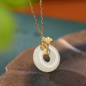 Hetian jade ginkgo leaf peace buckle necklace pendant on brown leaf tray, showing polished white nephrite jade with gold-plated ginkgo bail and delicate chain