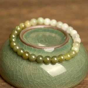 Two strands of gradient Hetian jade bracelets showcasing color variation on celadon ceramic bowl