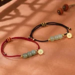 Two Hetian jade lotus bracelets with red and black woven cords and gold flower charms displayed on warm brown fabric background