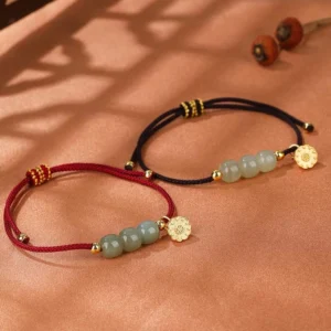 Two Hetian jade lotus bracelets with red and black woven cords and gold flower charms displayed on warm brown fabric background