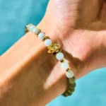 Hetian jade lotus abundance bracelet worn on left wrist, showing gradient white to green jade beads with gold lotus charm against blue fabric