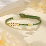 Hetian jade peace buckle bracelet with pale green jade and green braided cord, displayed on warm floral backdrop with gold accents