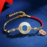 Hetian jade peace buckle bracelet with sterling silver chain, red sacred cord, and cubic zirconia accents on dark blue background - oblique top view