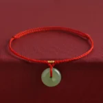 Red string Hetian jade peace buckle bracelet with gold accent knot, front view on dark fabric