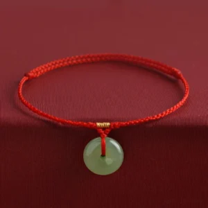 Red string Hetian jade peace buckle bracelet with gold accent knot, front view on dark fabric