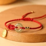 Hetian jade peace buckle bracelet with red string and fu character charm on wooden surface, three-quarter view showing jade donut centerpiece and gold metal accessories