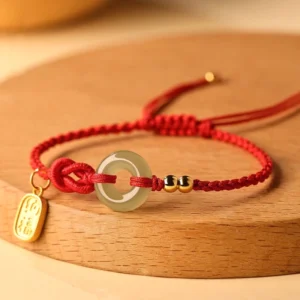 Hetian jade peace buckle bracelet with red string and fu character charm on wooden surface, three-quarter view showing jade donut centerpiece and gold metal accessories