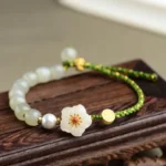 Hetian jade peach blossom bracelet with green woven cord and white floral charm, overhead flat lay on wooden stand