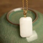 Hetian jade rectangular pendant necklace with gold metal chain, polished white jade close-up on ceramic teapot backdrop