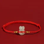Red string Hetian jade rabbit bracelet with gold accents, flat lay on red background
