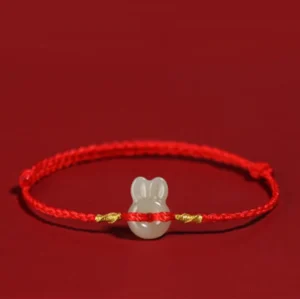Red string Hetian jade rabbit bracelet with gold accents, flat lay on red background