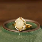 Natural Hetian jade adjustable ring with cubic zirconia halo and gold-plated copper band, front flat lay view on celadon ceramic