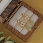 Hetian jade and tridacna stone flower drop earrings displayed in wooden jewelry box, overhead view showing gold-tone hardware and pale green jade bead