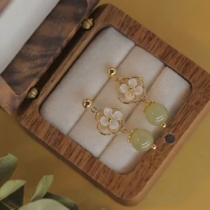 Hetian jade and tridacna stone flower drop earrings displayed in wooden jewelry box, overhead view showing gold-tone hardware and pale green jade bead