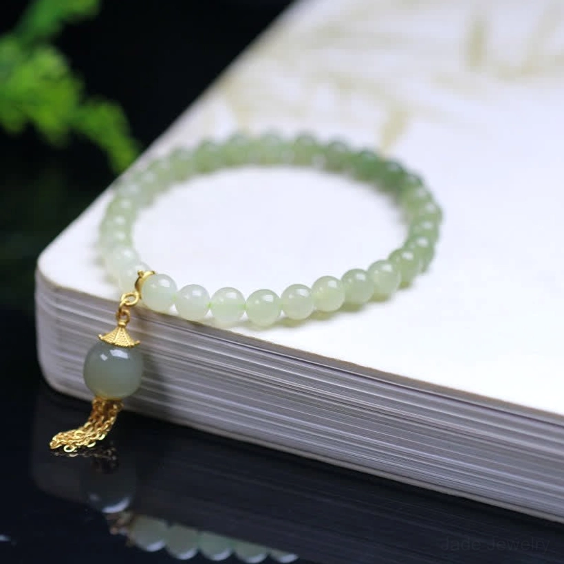 Hetian jade beaded bracelet flat lay on hardcover book, gradient green jade beads with gold tassel pendant on reflective tabletop