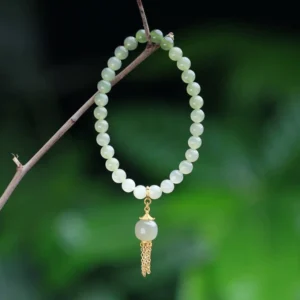 Hetian jade wealth bracelet with sterling silver charm, pale green jade beads with gold tassel pendant displayed against natural green plant background