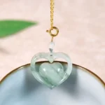 Hollow heart jade prosperity necklace with gold-plated chain displayed on light blue ceramic bowl, overhead close-up view showing intricate double heart design