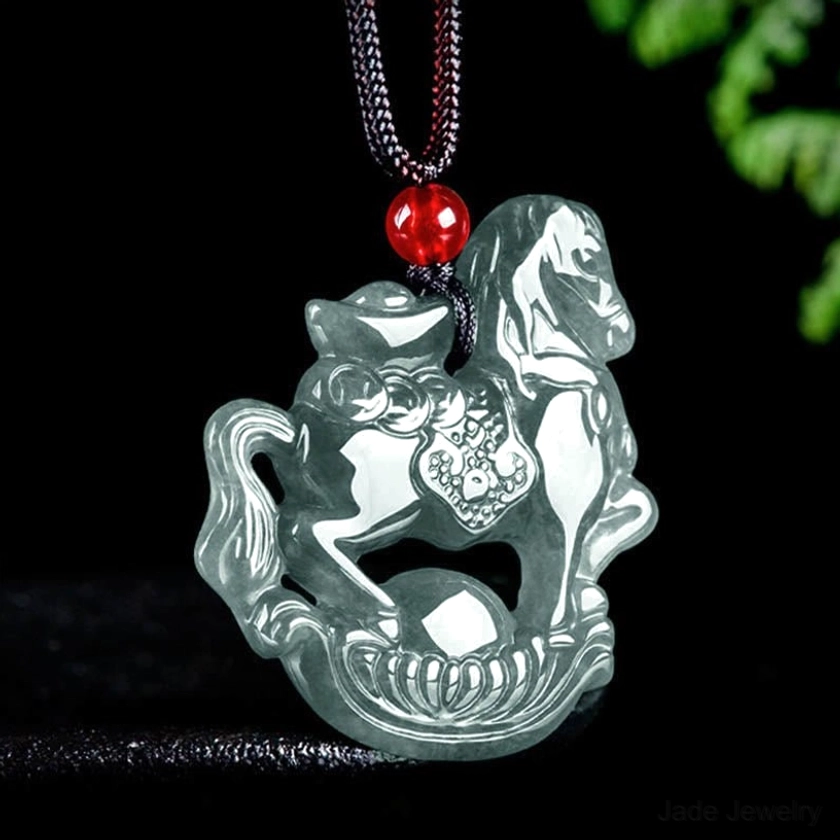 Close-up three-quarter view of hand-carved horse ingot jade pendant showing detailed engraving and smooth polished surface with red accent bead and black hanging cord on reflective studio surface