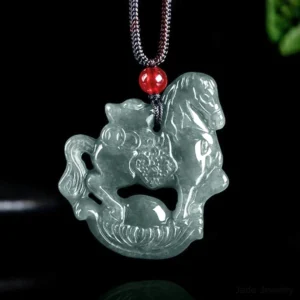 Horse zodiac jade pendant necklace with intricate carved horse and ingot design on translucent grey jade, featuring red accent bead and dark braided cord against dark studio background with green foliage