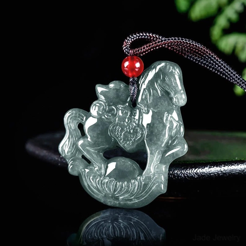 Side angled view of horse zodiac jade pendant necklace displayed on dark reflective stand showing carving depth, grey jade coloring, and dark fabric cord with red bead accent