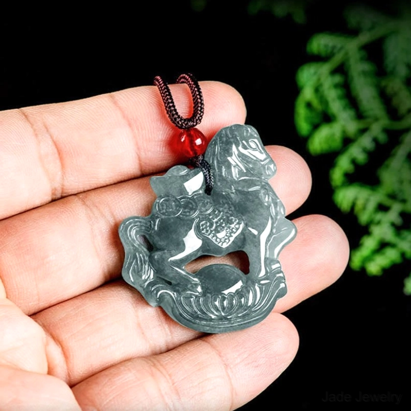 First-person hand-held view of horse zodiac jade pendant necklace held in human palm against green foliage background showing real-life size and translucent grey-green jade coloring