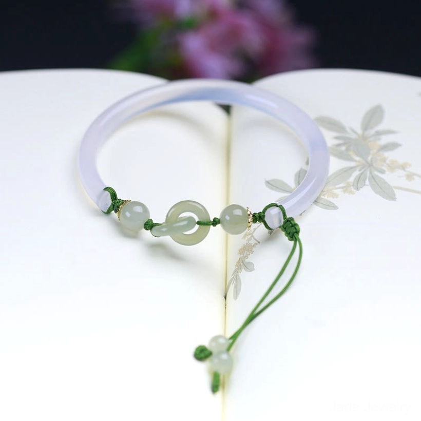 Purple-white Hetian jade bangle bracelet with interlocking charm pendant and green cord, flat lay on open floral print book