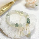 Natural gradient jade beaded charm bracelet with two-tone green and white jade beads, gold-tone pendants, displayed on an ornate golden tray with decorative coral and flowers for prosperity and abundance