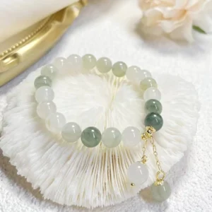 Natural gradient jade beaded charm bracelet with two-tone green and white jade beads, gold-tone pendants, displayed on an ornate golden tray with decorative coral and flowers for prosperity and abundance