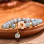 Jade abundance flower bracelet flat lay top view featuring gray-blue jade beads, white floral charm with red gem center, and gold spacers on wooden surface