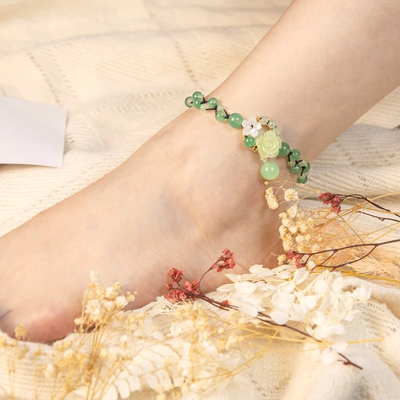 Detailed close-up of jade ankle bracelet with floral charms and beads on foot with dried wild flowers and linen