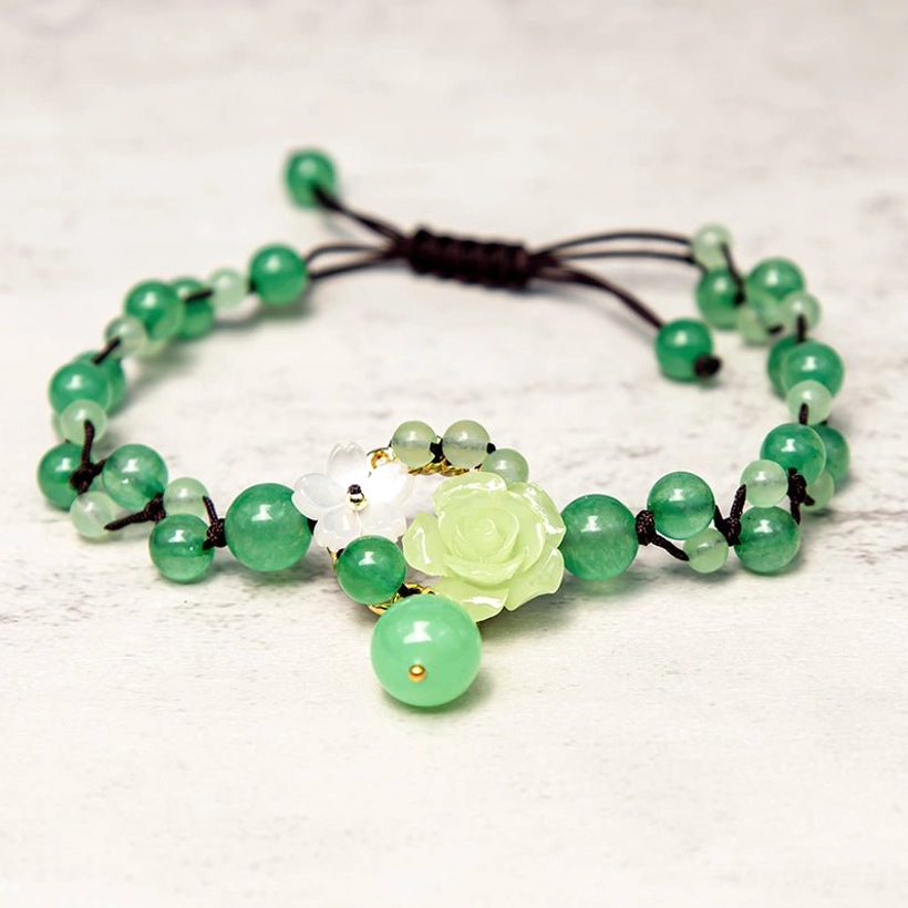 Top-down flat lay of jade braided anklet with green beads, black cord, and rose and flower charm accents on marble surface