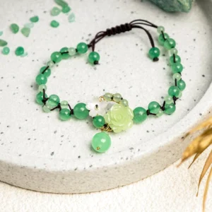 Overhead view of jade braided ankle bracelet featuring green beads, woven cord, and rose flower charm on scattered jade chips