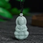 Pale green jade Avalokitesvara Guanyin pendant necklace with detailed carving, front view displayed on dark stone with botanical background