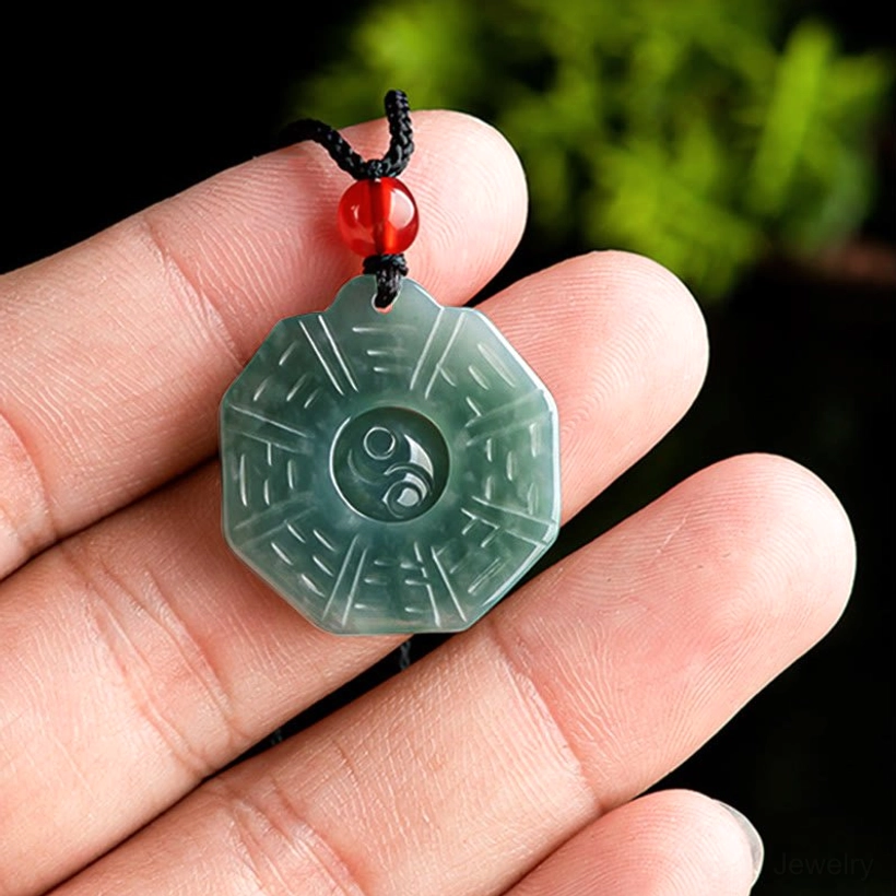 Mini jade Bagua pendant held in human hand on plain background showing compact size with red bead on black cord