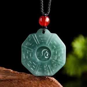 Natural jade Bagua Yin Yang pendant necklace with dark green carved jade, red agate bead, and black cord displayed on wooden base with green foliage