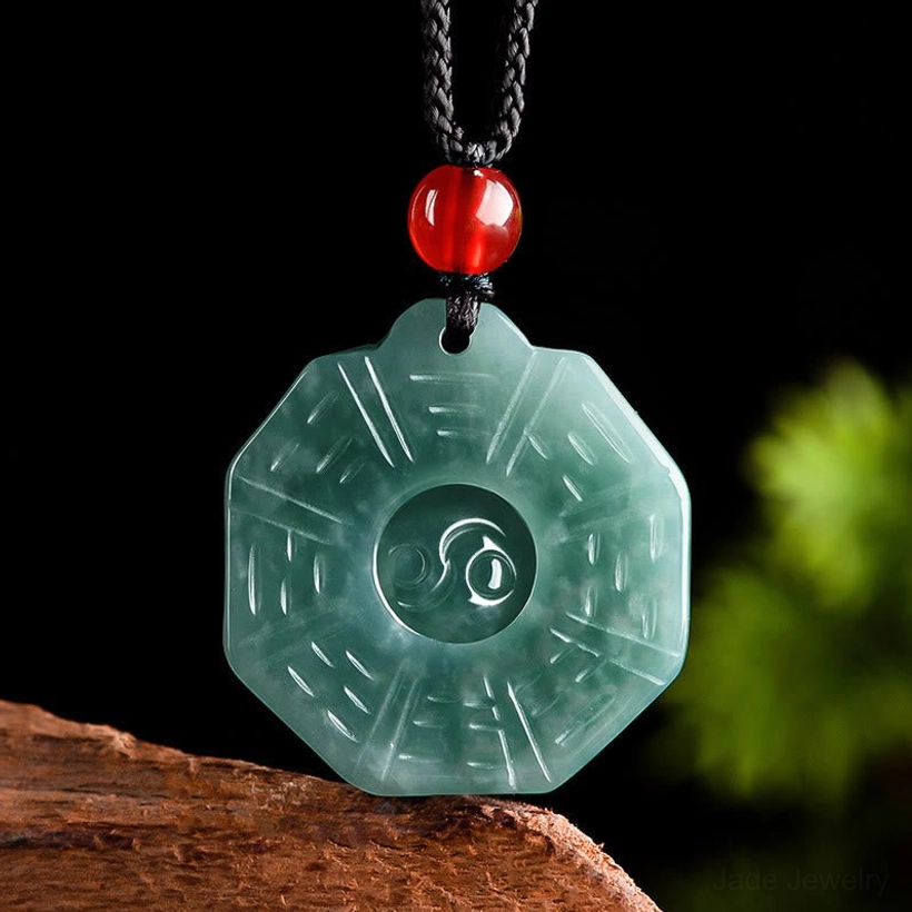 Natural jade Bagua Yin Yang pendant necklace with dark green carved jade, red agate bead, and black cord displayed on wooden base with green foliage